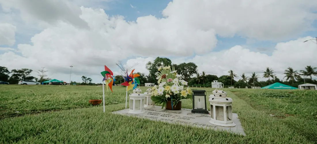 A grassy outdoor memorial with white lanterns, a flower arrangement, and colorful pinwheels on a sunny day under a blue sky.