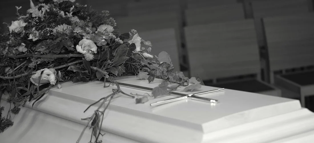 Black-and-white photo of a closed coffin with a floral arrangement resting on top at a funeral.