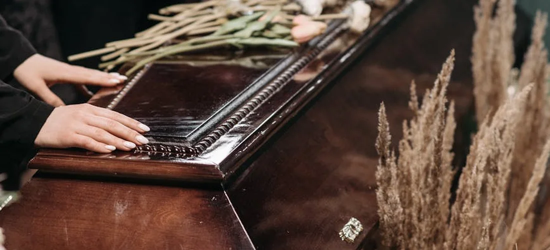 Close-up of a coffin with hands placed near it, with dried flowers nearby, illustrating planning for a postponed funeral
