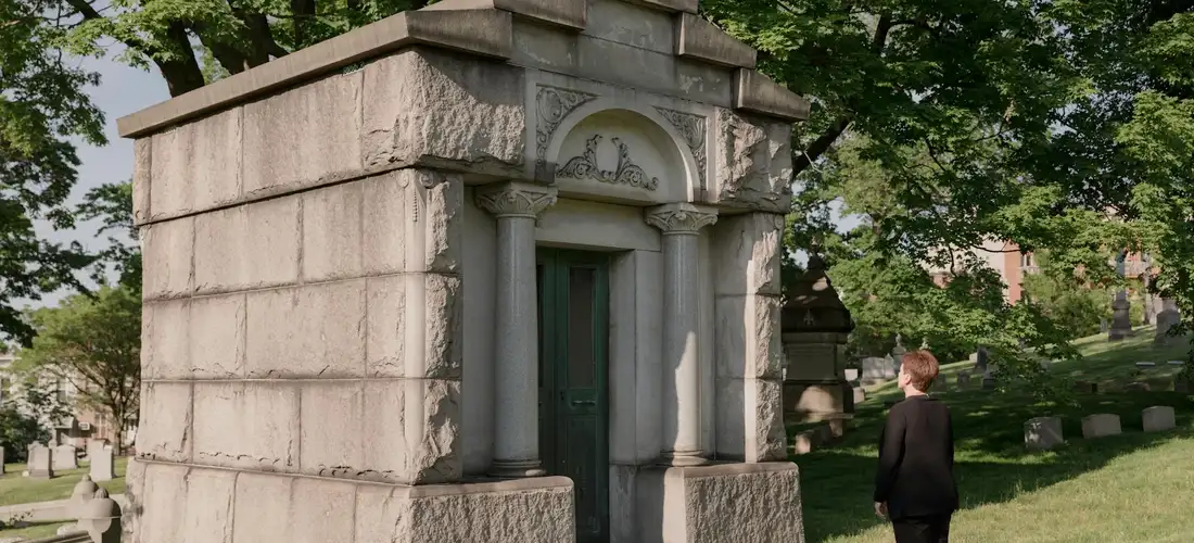 A person in a dark suit stands near a stone mausoleum in a quiet cemetery, conveying a solemn setting.