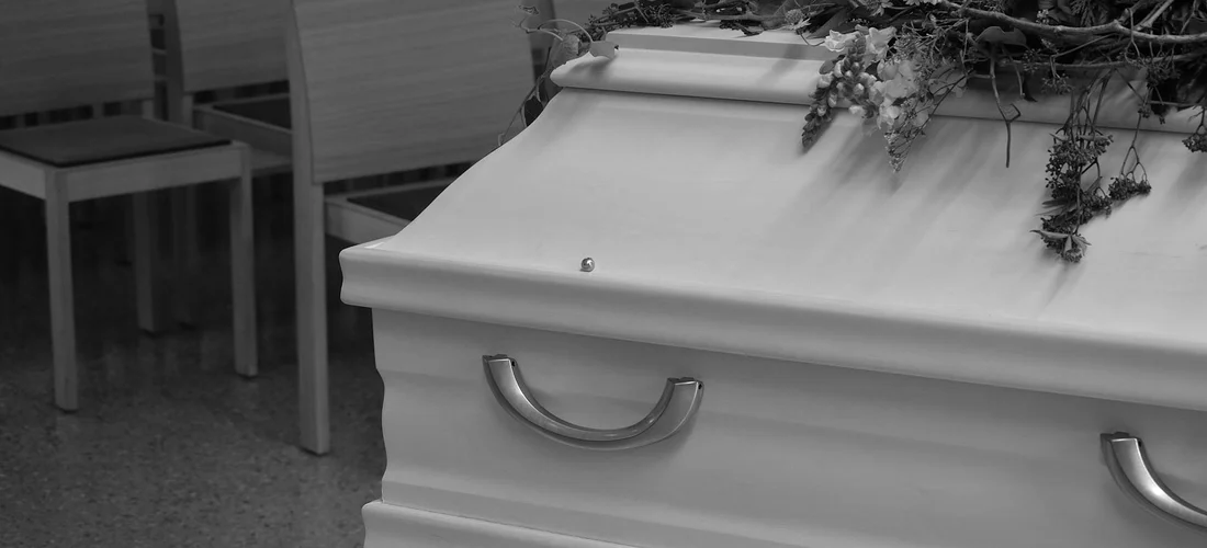 Black-and-white photograph of a closed coffin with a floral arrangement on top in a funeral setting.