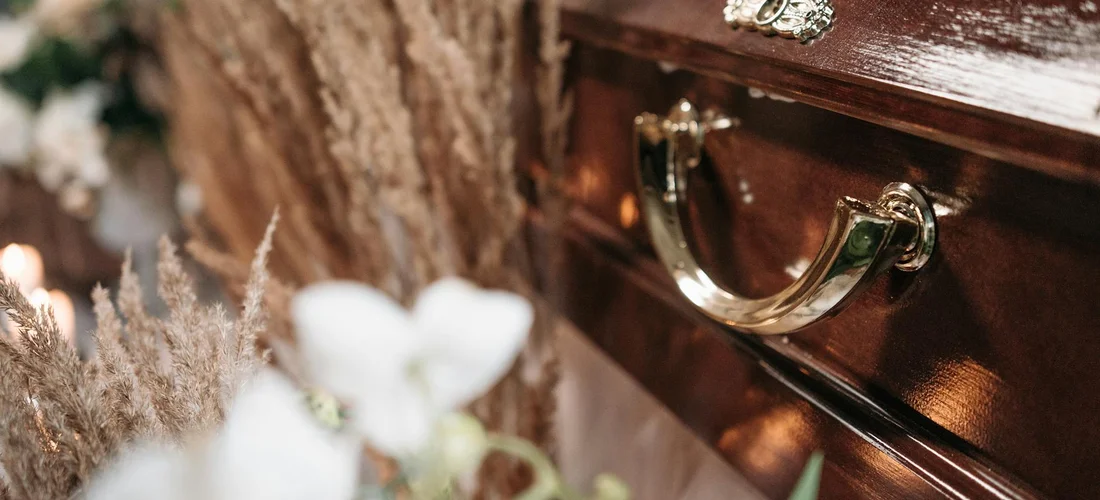 Close-up of a polished wooden coffin with a brass handle, with blurred white flowers in the foreground.