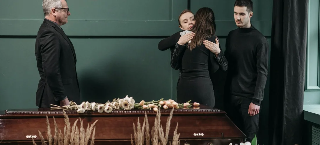 A group of mourners embrace near a closed casket, with floral arrangements on top, in a funeral setting.