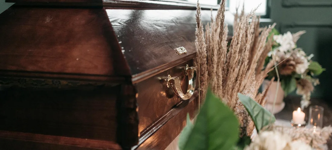 Close-up of a dark wooden coffin in a funeral setting with floral arrangements.