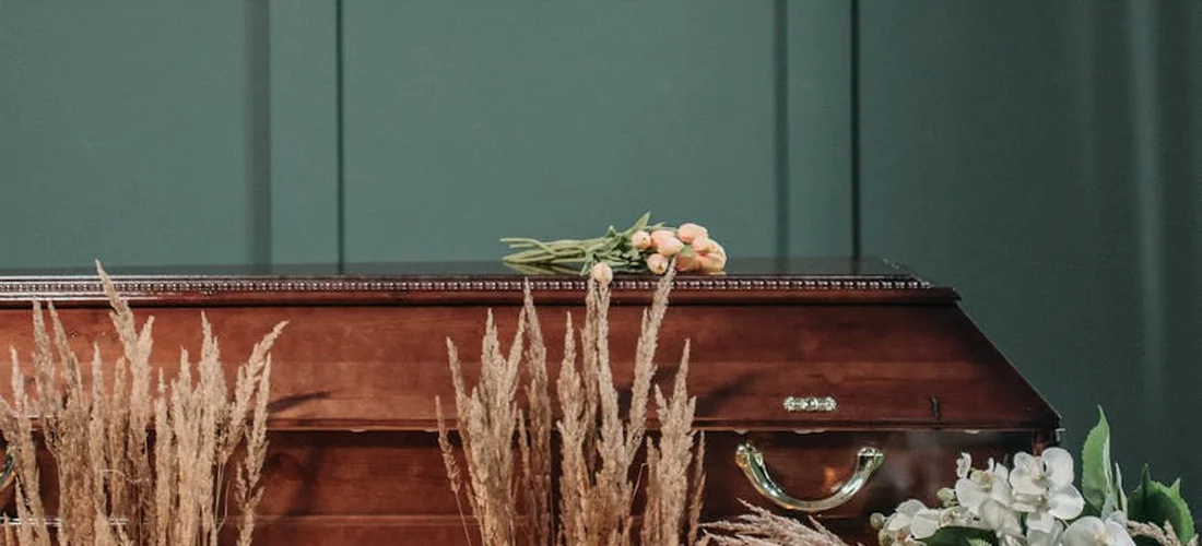 Closed wooden coffin adorned with flowers and dried grasses in front of a dark green backdrop.