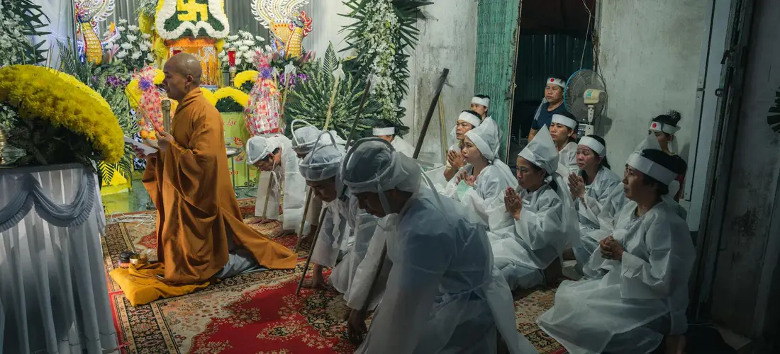 A Buddhist funeral ceremony with a monk in orange robes leading attendees dressed in white robes and head coverings, seated in prayer inside a temple with floral decorations.