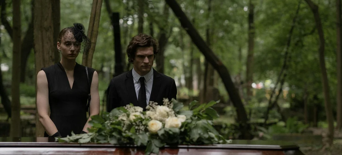 Two mourners, a man in a suit and a woman in a black dress, stand beside a casket adorned with white flowers in a wooded cemetery, looking solemn.