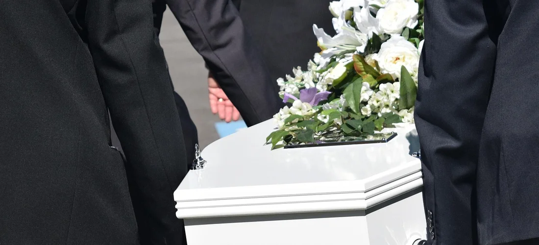Mourners in dark suits stand beside a white casket decorated with flowers.