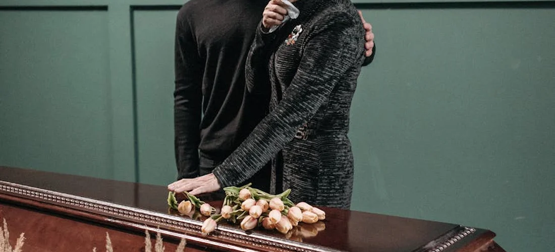 Two mourners in dark clothing stand beside a polished casket adorned with a small bouquet of pale pink roses in a funeral home.