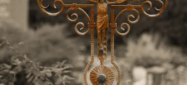 Black-and-white photograph of an ornate iron crucifix with a figure of Jesus, set in a blurred cemetery garden.