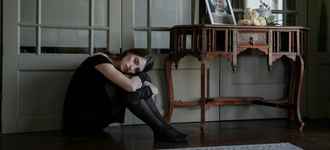 A person sits on the floor with knees drawn up, resting their head on their arms, beside a wooden console table with a framed photograph and flowers, conveying contemplation and sorrow.