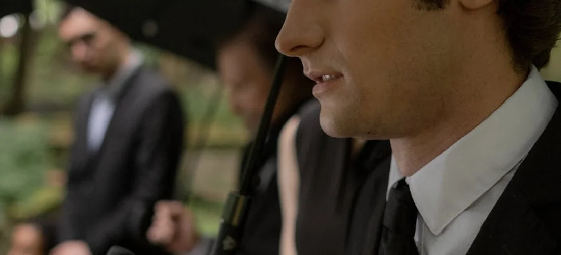Close-up of a man in a suit at a funeral, with other attendees blurred in the background.
