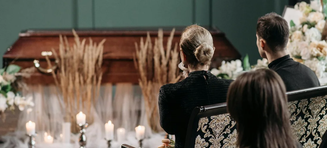 People seated in a funeral parlor facing a casket with lit candles and floral arrangements.