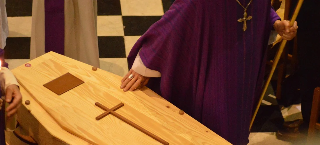 A wooden coffin with a cross on its lid rests on a table in a chapel. A clergyman wearing purple vestments stands nearby.