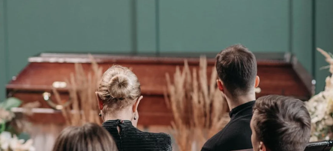 Attendees at a funeral viewed from behind, with a piano in the background, suggesting a somber service and the moment to write a condolence message from colleagues.