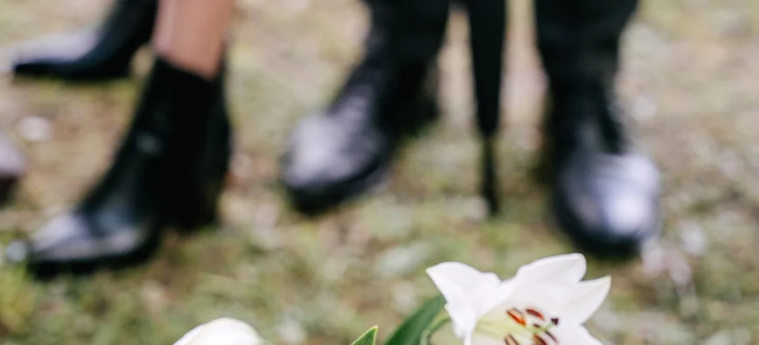 Close-up of a white flower in sharp focus with blurred mourners' feet and shoes in dark clothing in the background, suggesting a funeral setting.