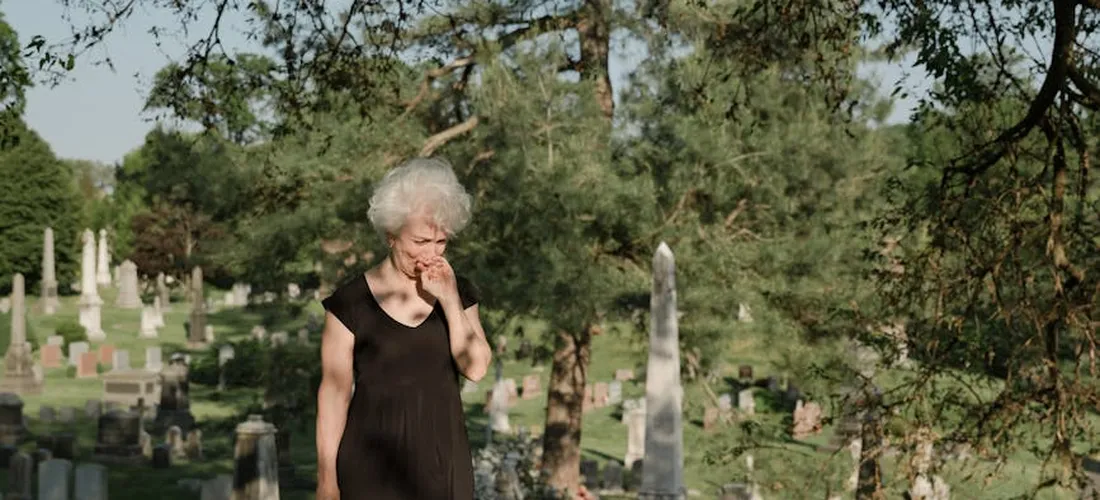 An elderly woman in a black dress stands among gravestones in a cemetery, appearing to mourn.