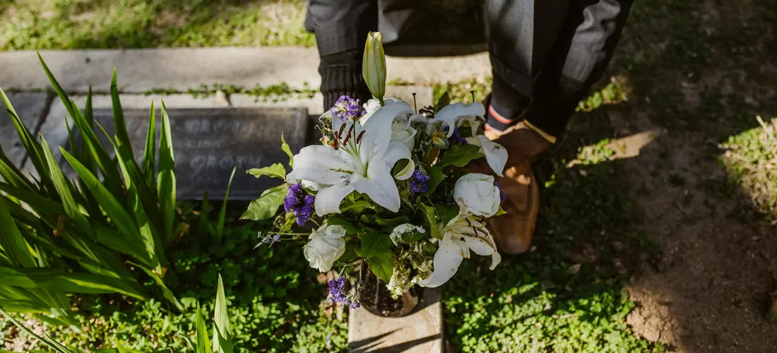 A person places a bouquet of white lilies and purple flowers on a grave, with greenery and a headstone nearby.