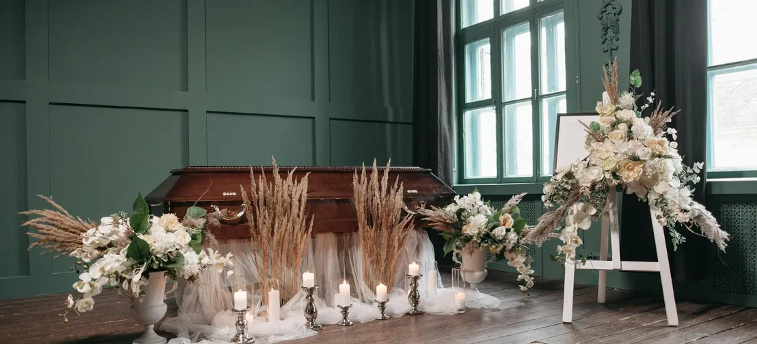 A funeral setting featuring a wooden casket, white floral arrangements, and candlelit accents in a room with green walls, illustrating traditional floral displays.