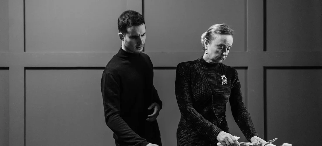 Black-and-white photograph of a man and a woman in formal attire standing near a table, conveying a somber mood typical of a funeral or memorial setting.