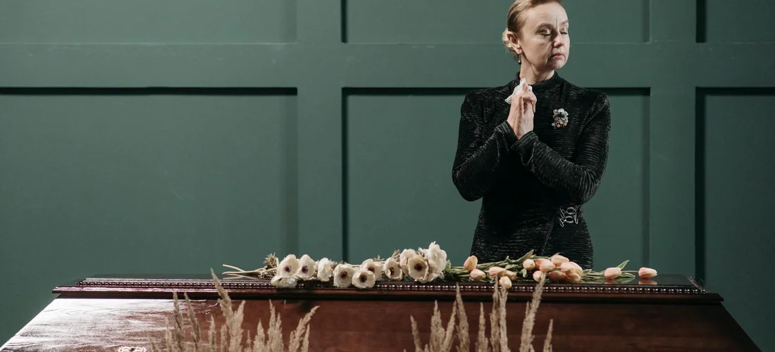 A person in a dark outfit stands beside a closed casket adorned with flowers, hands clasped in a prayer-like pose.