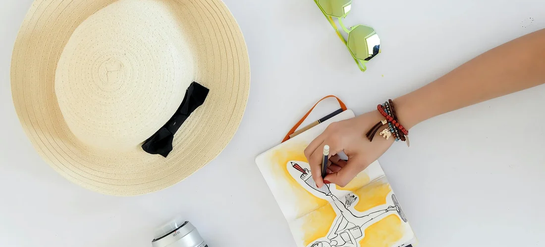 Overhead view of a light straw hat with a black ribbon beside a person’s hand sketching on a small notebook; a bracelet-wrapped wrist and a plant are visible on a bright surface.