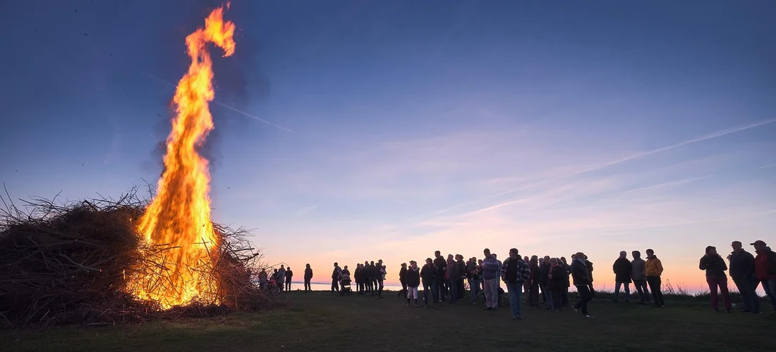 Silhouettes of mourners standing around a large cremation pyre at sunset.