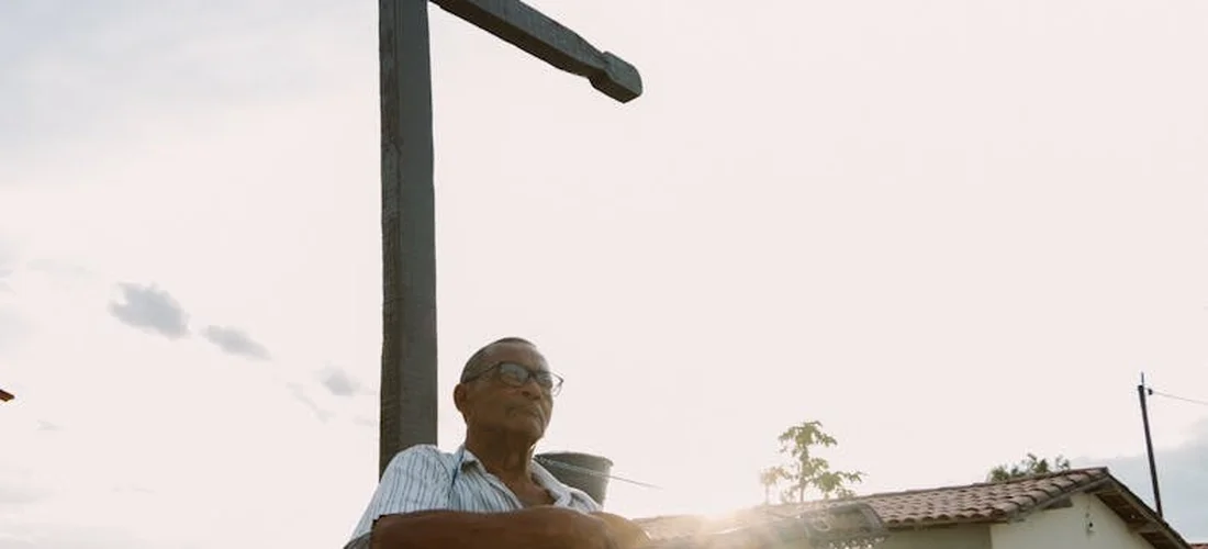 An older man wearing glasses stands outdoors beside a tall cross, with a bright sky in the background, suggesting reflection and remembrance associated with funeral services.