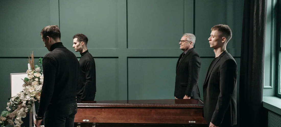 Four men in dark suits stand beside a coffin in a funeral setting, with minimal visible jewelry.