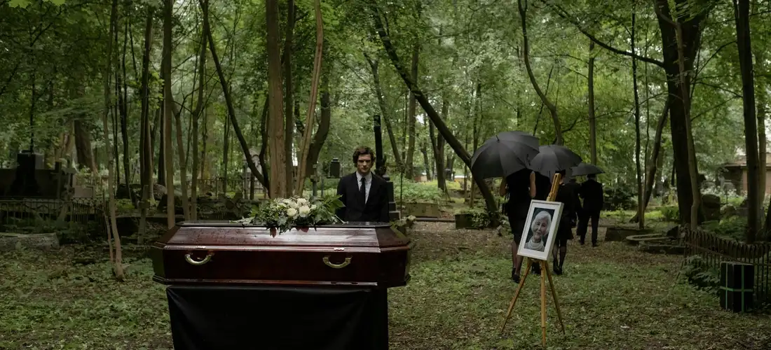A funeral scene in a wooded cemetery: a dark coffin sits at center with floral arrangements, a suited attendant stands nearby, and an easel holds a framed photograph under a black umbrella.