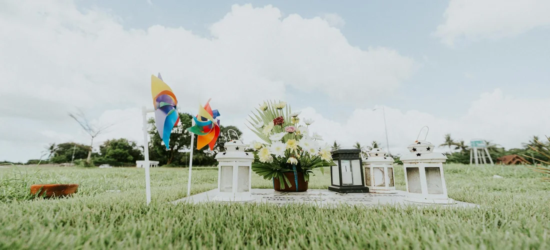 Outdoor memorial setup on a grassy field with white pedestals, flowers, and colorful flags under a blue sky