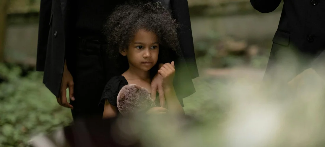 A young girl stands with adults dressed in black at a funeral, holding a stuffed toy.