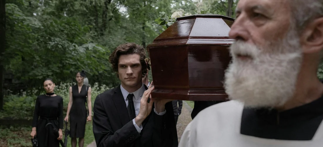 An outdoor funeral scene with pallbearers in black suits carrying a coffin; a bearded clergyman stands in the foreground.