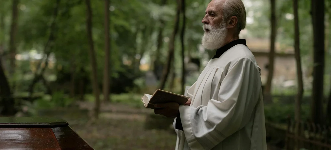 An elderly clergyman in a white robe stands outdoors in a wooded setting, holding a small book as he prepares for a service.