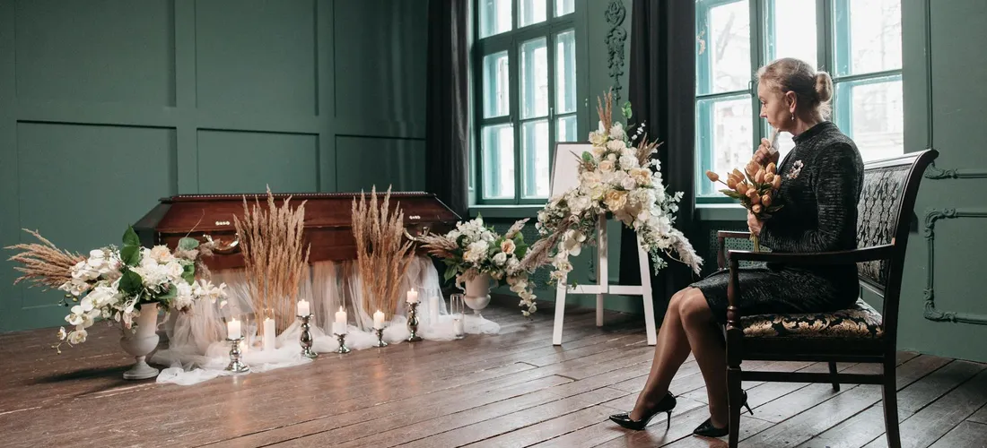 A woman seated in a quiet funeral space with a casket and floral arrangements, preparing to lead the ceremony.