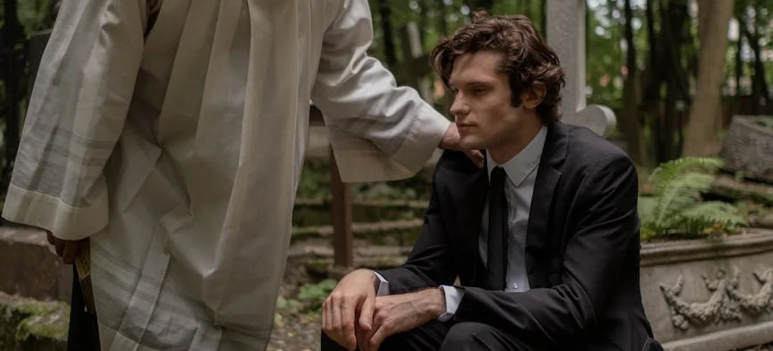 A man in a dark suit sits on a bench in a cemetery as another person wearing a light-colored outfit stands nearby, suggesting a planning discussion for a funeral service.