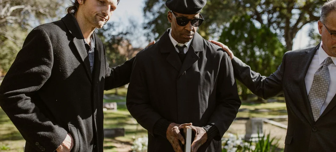 Three pallbearers in dark suits stand near a grave in a cemetery.