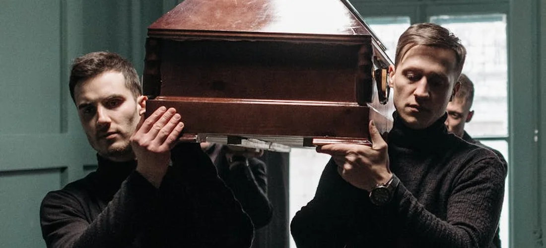 Two pallbearers in dark clothing carry a closed coffin, ready for a funeral service.