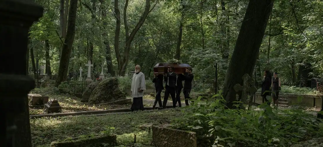 Funeral procession in a wooded cemetery with pallbearers carrying a casket