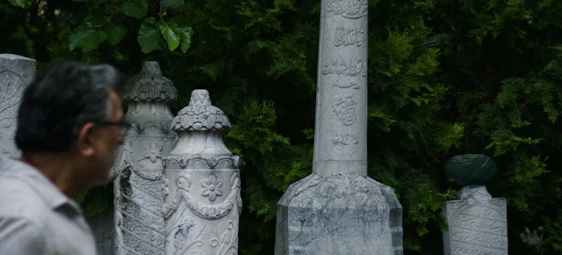 An older man wearing glasses stands beside weathered tombstones in a cemetery, suggesting the topic of funeral planning and related paperwork.