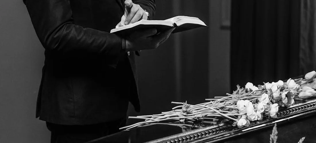 Person in a dark suit reading from an open book beside a flower-adorned casket at a funeral.