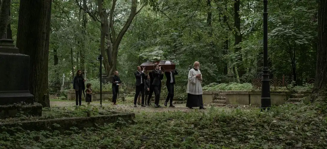 A funeral procession in a wooded cemetery park; pallbearers carry a coffin along a path while a mourner walks ahead.