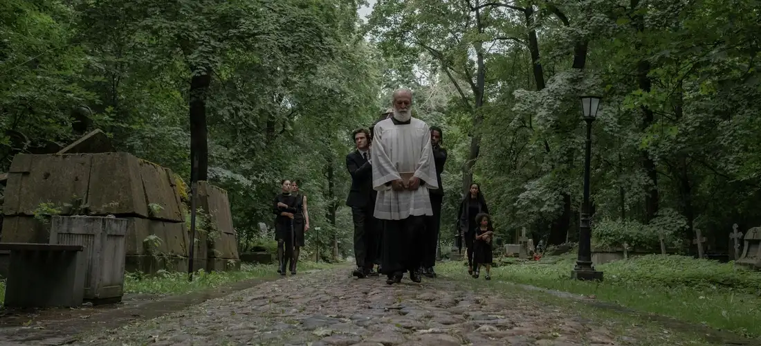 Clergy leading a funeral procession along a cobblestone path in a green park cemetery, with attendees walking behind.