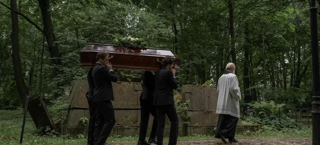 Pallbearers in dark suits carry a casket along a tree-lined cemetery path, followed by a person in a light-colored coat.