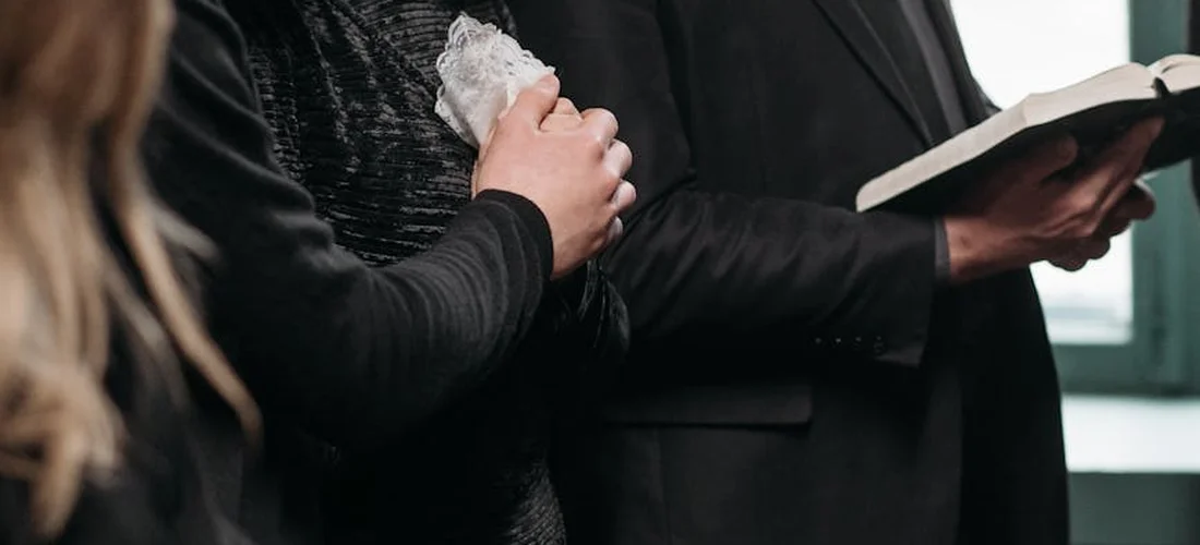 Mourners dressed in black at a funeral service, with one person holding a booklet or program, conveying the human context of end-of-life rites