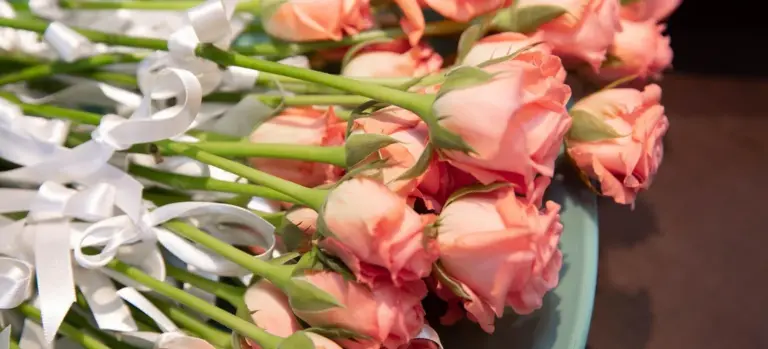 Bouquet of pink roses with white ribbons arranged on a surface, suggesting a funeral or memorial setting.