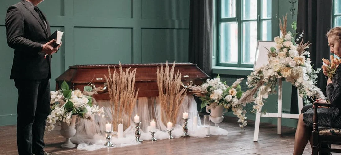 A man in a dark suit stands with a book near a casket in a chapel, surrounded by white flowers, candles, and guests seated, illustrating a solemn funeral scene.
