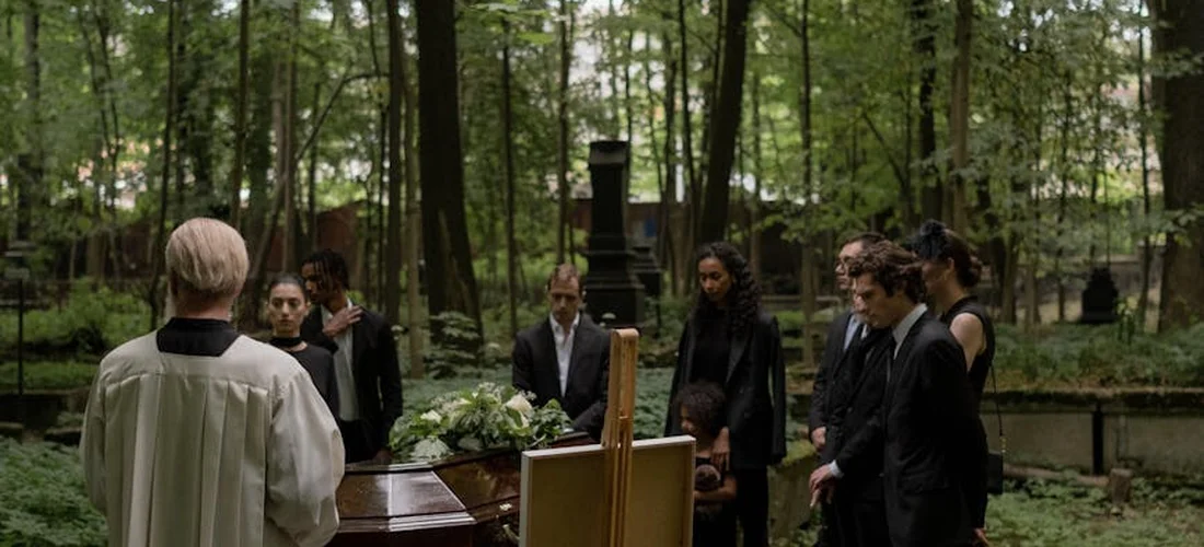 Mourners in dark formal attire gather around a coffin in a serene forest setting during a funeral service, with an officiant at the front.