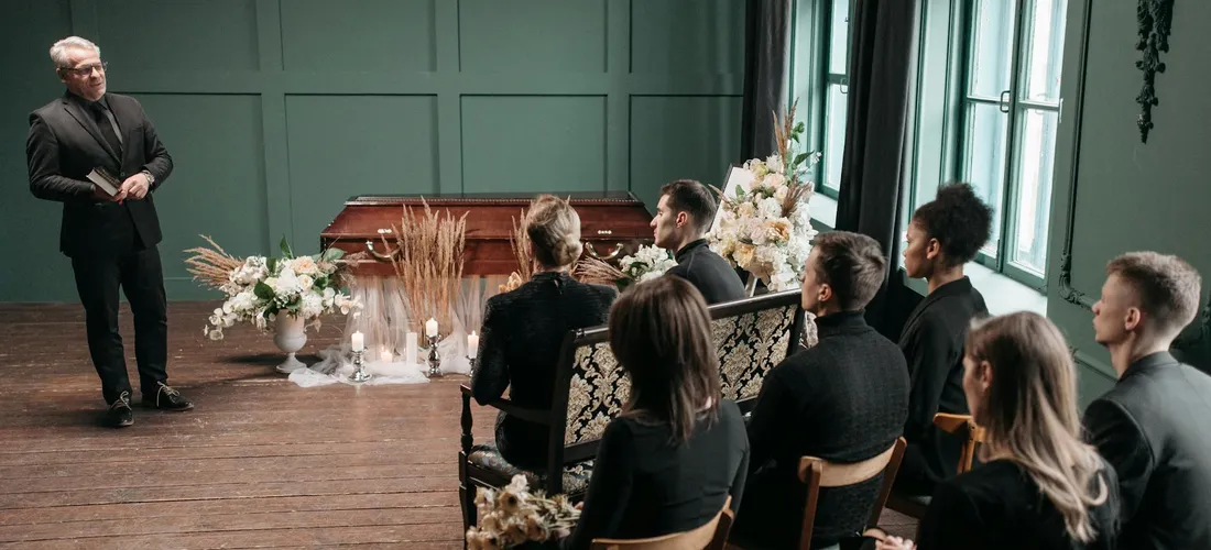 A speaker addresses mourners seated in a funeral service setting, with dark attire and floral arrangements in a softly lit room.