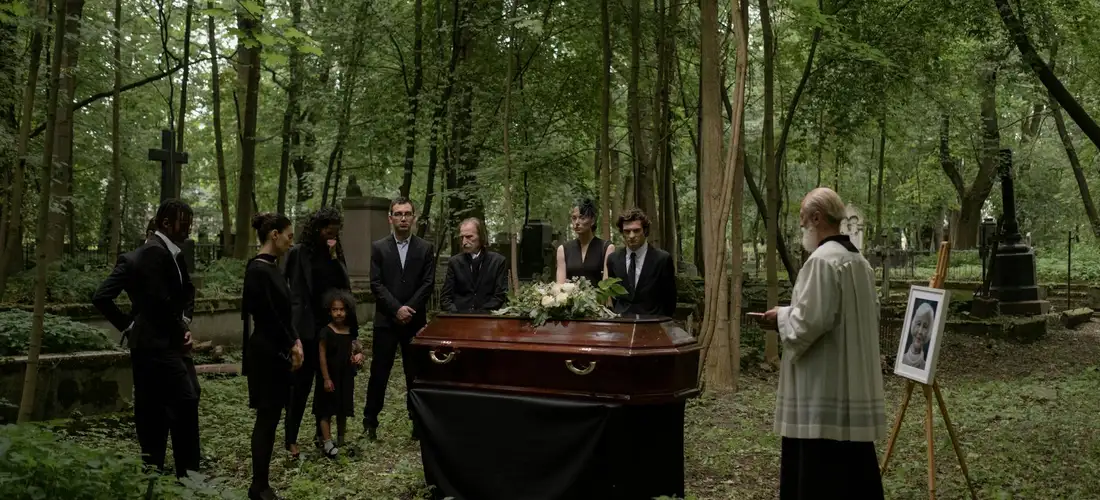 In a forested cemetery, pallbearers in dark suits stand around a casket positioned on a bier surrounded by mourners; an officiant with a light-colored robe speaks, while a framed portrait rests on an easel beside the grave.